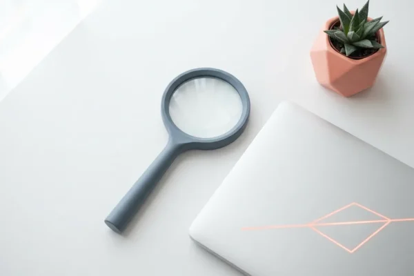 Vergrootglas op wit bureau naast laptop en kleine vetplant, minimalistische flatlay in wit, koraal en leisteengrijs.